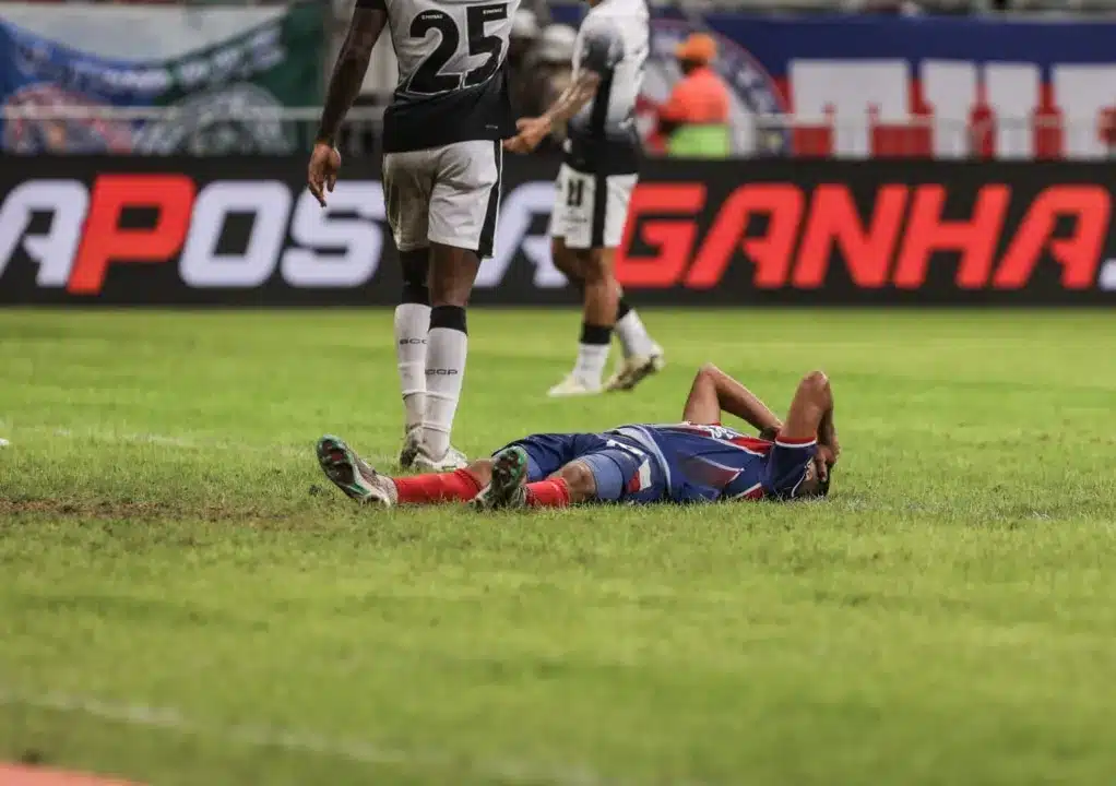 Jogador do Bahia está deitado no gramado com as mãos na cabeça. Ao fundo, dois jogadores do Corinthians caminham.