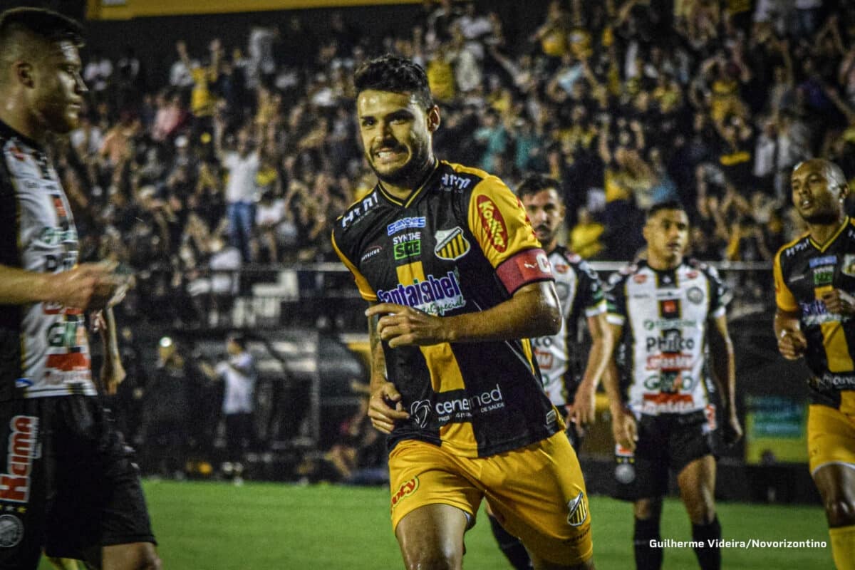 Jogador do Novorizontino comemora um gol. Ao fundo, jogadores do Operário estão desanimados por terem sofrido um gol.