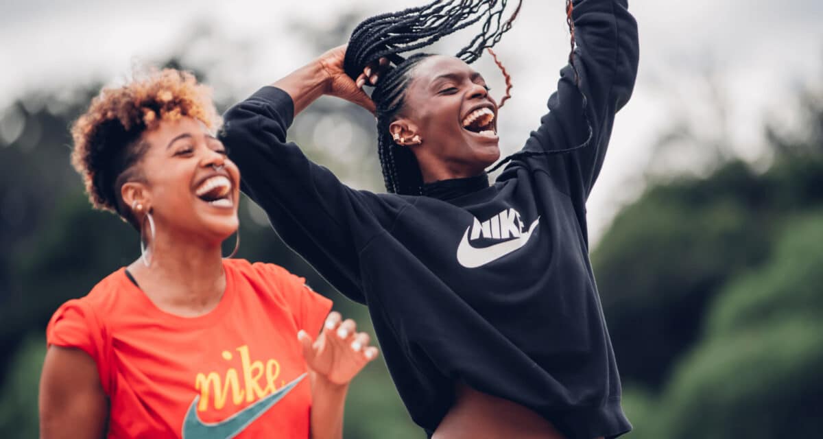 Duas mulheres negras riem largamente. Ambas estão vestindo camisa da marca esportiva Nike.