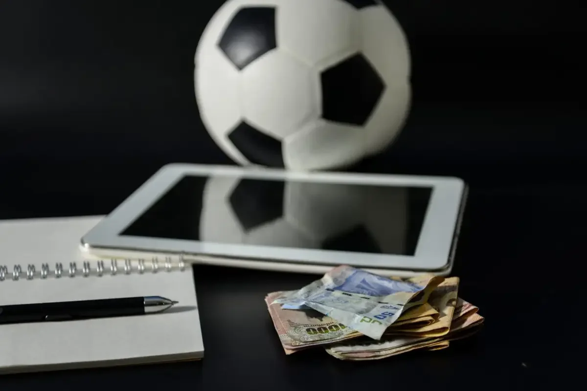 Uma bola de futebol, um tablet, um cadernos e notas de dinheiro estão dispostos numa mesa preta.