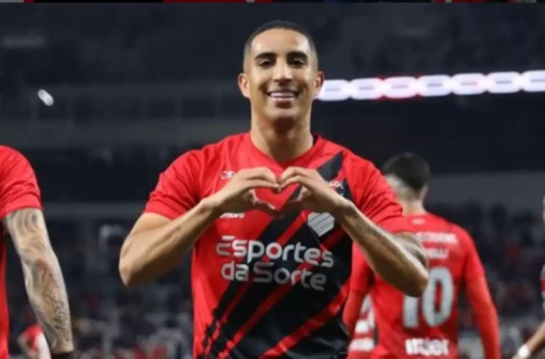 Jogador do Athletico-PR sorridente comemora gol fazendo sinal de coração com as mãos para a câmera