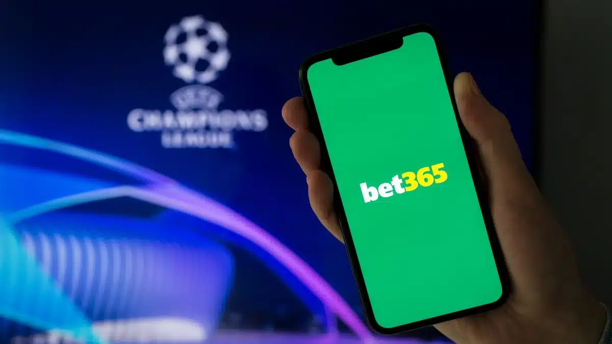 Um celular está em destaque nesta imagem. Na tela do telefone está a logo da bet365. Atrás do celular está a logomarca da Liga dos Campeões.