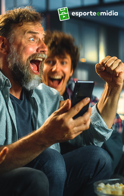 Foto de homem branco comemorando ganhos com celular na mão. Um homem no fundo comemora sorrindo. Logo do Esporte e Mídia aparece no canto superior direito da imagem.
